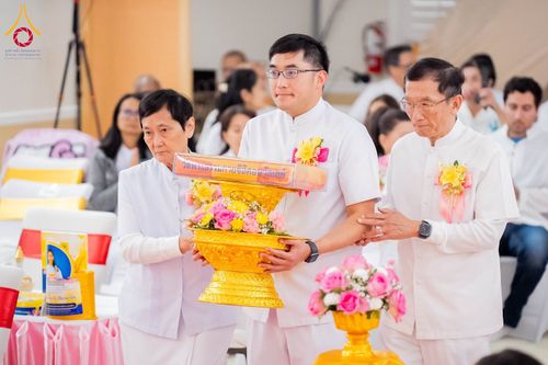 กลุ่มคนในชุดสีขาวกำลังถือพานพุ่มดอกไม้และผ้าไตรในพิธีทางศาสนา