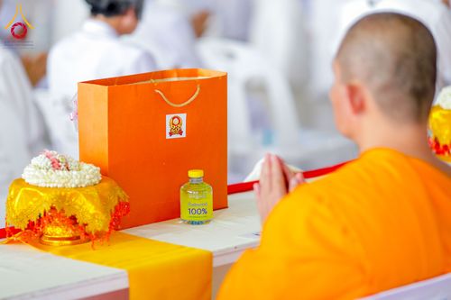 พระภิกษุพนมมือไหว้หน้าโต๊ะที่มีถุงกระดาษสีส้มและพานพุ่มดอกไม้