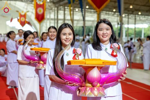 หญิงสาวสองคนยิ้มแย้มถือพานพุ่มประดับผ้าไตรจีวรสีเหลืองทองในพิธีทางศาสนา