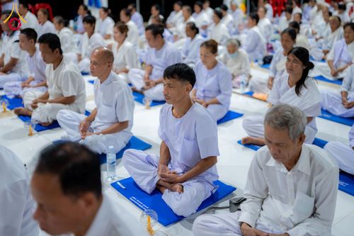 กลุ่มคนสวมชุดสีขาวนั่งขัดสมาธิบนเบาะรองนั่งสีน้ำเงินเพื่อปฏิบัติธรรมภายในอาคาร