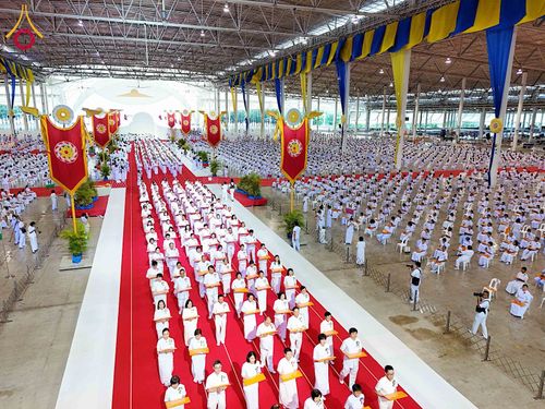 พุทธศาสนิกชนชุดขาวเดินแถวถือพานผ้าป่าบนพรมแดงในอาคารขนาดใหญ่ที่มีผู้คนนั่งร่วมพิธีจำนวนมาก