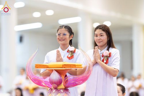 หญิงสองคนสวมชุดสีขาวพนมมือประกอบพิธีทางศาสนา โดยมีพานวางผ้าไตรสีเหลืองทองอยู่ด้านหน้า