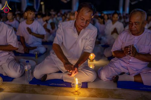 ชายสูงวัยในชุดสีขาวกำลังจุดเทียนในโคมแก้วขณะนั่งร่วมพิธีกรรมกับกลุ่มผู้ปฏิบัติธรรม