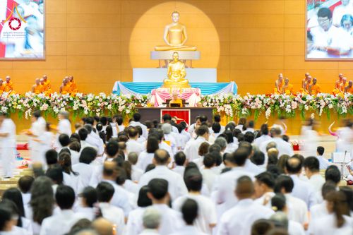 พุทธศาสนิกชนสวมชุดขาวนั่งร่วมพิธีทอดผ้าป่าสมทบกฐินภายในสภาธรรมกายสากลที่มีพระภิกษุสงฆ์และพระพุทธรูปบนเวที