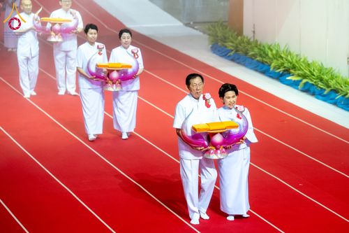 กลุ่มคนในชุดขาวเดินถือพานพุ่มผ้าไตรบนพรมแดงในพิธีทางศาสนา