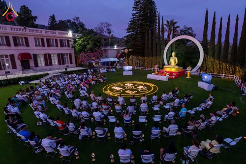กลุ่มคนนั่งสมาธิบนสนามหญ้าหน้าพระพุทธรูปและวงกลมประทีปในบรรยากาศยามเย็น