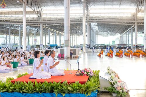 พุทธศาสนิกชนชุดขาวนั่งพนมมือบนเวทีในอาคารสภาธรรมกายสากล โดยมีพระภิกษุสงฆ์นั่งอยู่ด้านหลัง