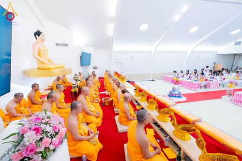 พระภิกษุสงฆ์ห่มจีวรสีเหลืองทองนั่งเรียงแถวประกอบพิธีทางศาสนาภายในอาคารที่มีพระพุทธรูปประดิษฐานอยู่ด้านหน้า