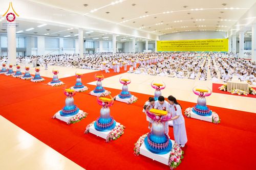 พุทธศาสนิกชนชุดขาวร่วมพิธีทอดผ้าป่าสมทบกฐินภายในอาคารขนาดใหญ่ โดยมีพานพุ่มผ้าป่าวางบนฐานดอกไม้เรียงรายบนพรมสีแดง