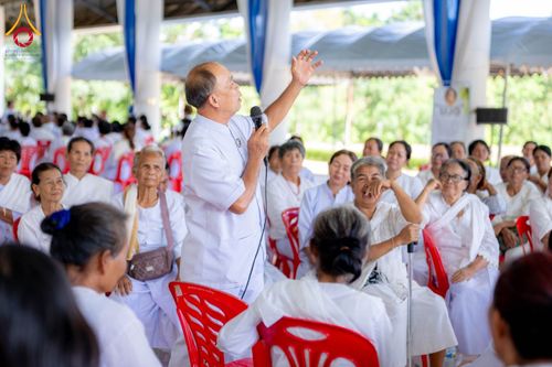 วิทยากรชายยืนพูดบรรยายหน้ากลุ่มผู้เข้าอบรมที่สวมชุดสีขาวนั่งอยู่บนเก้าอี้พลาสติกสีแดงภายในอาคาร