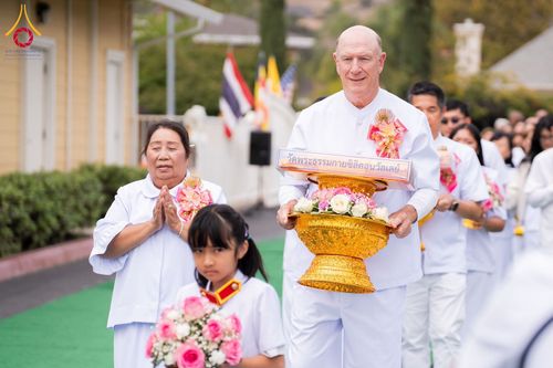 ชายชาวต่างชาติและหญิงไทยแต่งกายชุดขาวกำลังถือพานพุ่มดอกไม้และป้ายชื่อวัดเดินในขบวน