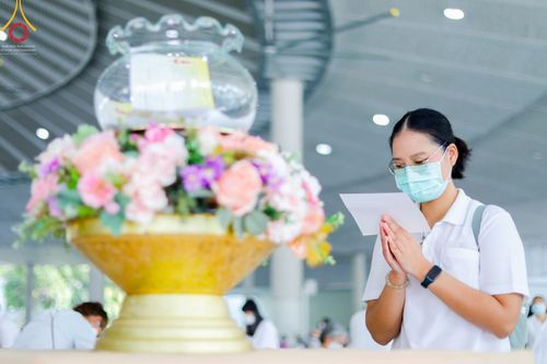 หญิงสาวสวมหน้ากากอนามัยและแว่นตาสวดมนต์และถือกระดาษอยู่ในมือ หน้าพานพุ่มดอกไม้ในพิธีทางศาสนาภายในอาคาร