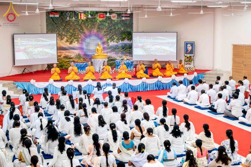 พระภิกษุสงฆ์และพุทธศาสนิกชนร่วมพิธีทางศาสนาภายในอาคารที่มีฉากหลังเป็นภาพต้นไม้และพระพุทธรูป