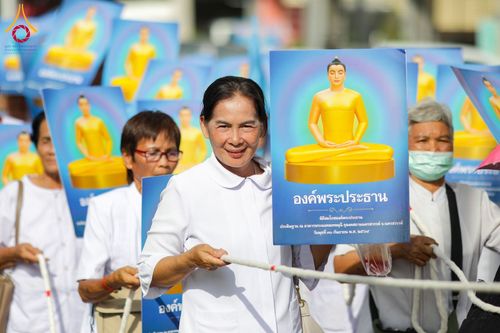 กลุ่มคนสวมชุดขาวถือป้ายภาพพระพุทธรูปสีทองและจับเชือกในขบวนแห่
