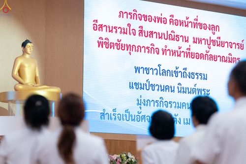 กลุ่มคนสวมชุดขาวหันหลังให้กล้องมองไปยังจอโปรเจกเตอร์ที่มีข้อความภาษาไทยและพระพุทธรูปสีทอง