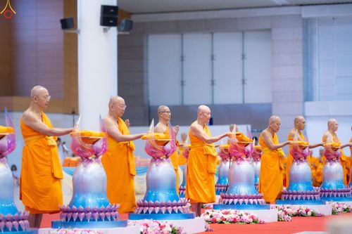 พระภิกษุสงฆ์ในจีวรสีส้มกำลังประกอบพิธีทอดผ้าป่าโดยวางผ้าไตรบนพานพุ่มดอกบัวจำลองภายในอาคารขนาดใหญ่