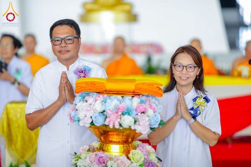 ชายและหญิงสวมชุดขาวประนมมือไหว้หน้าพานพุ่มดอกไม้และผ้าไตรจีวรในพิธีทางศาสนา