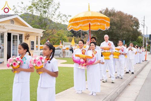 กลุ่มคนในชุดขาวเดินแถวถือพานดอกไม้และผ้าไตรในพิธีทางศาสนาพุทธ