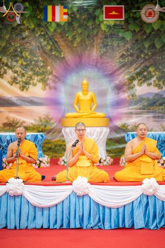 พระสงฆ์สามรูปนั่งพนมมือบนเวทีที่มีฉากหลังเป็นภาพพระพุทธรูปและต้นโพธิ์