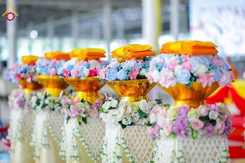 พานพุ่มสีทองประดับดอกไม้และผ้าไตรวางเรียงกันหลายใบในพิธีทางศาสนา