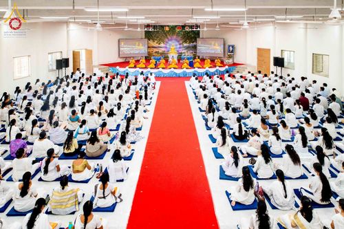 พุทธศาสนิกชนสวมชุดสีขาวนั่งร่วมพิธีในหอประชุมขนาดใหญ่โดยมีพระภิกษุสงฆ์อยู่บนเวทีด้านหน้า