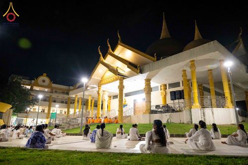 กลุ่มคนสวมชุดขาวนั่งปฏิบัติธรรมบนลานหน้าอาคารทรงไทยในยามค่ำคืน