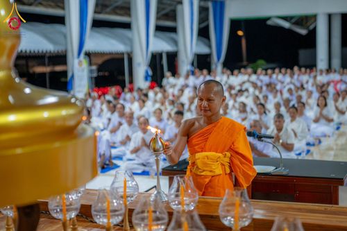 พระสงฆ์กำลังจุดประทีปในพิธี โดยมีผู้เข้าร่วมโครงการอบรมนั่งพนมมืออยู่เบื้องหลัง