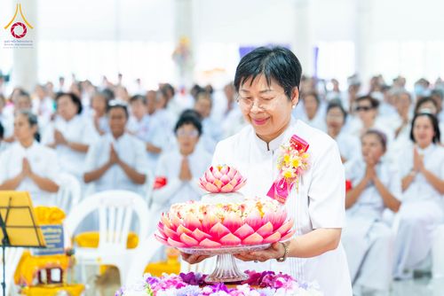 หญิงไทยสวมชุดขาวถือพานดอกบัวสีชมพูในพิธีทางศาสนา โดยมีผู้เข้าร่วมพิธีสวมชุดขาวนั่งอยู่ด้านหลัง