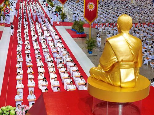 พุทธศาสนิกชนชุดขาวนั่งเรียงแถวบนพรมแดงในพิธีทอดผ้าป่า โดยมีรูปปั้นทองคำตั้งอยู่ด้านหน้า
