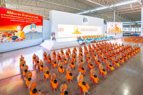 พระภิกษุสงฆ์จำนวนมากนั่งเรียงแถวบนเก้าอี้สีแดงภายในอาคารเพื่อเตรียมประกอบพิธีตักบาตร