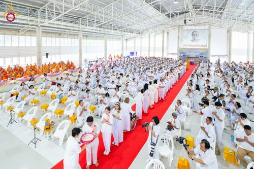 พุทธศาสนิกชนจำนวนมากสวมชุดขาวร่วมพิธีทางศาสนาภายในอาคารขนาดใหญ่ มีการเดินแถวบนพรมแดงและนั่งเก้าอี้สีขาวอย่างเป็นระเบียบ
