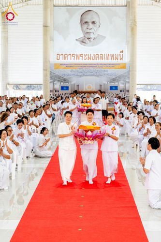 กลุ่มคนสวมชุดขาวเดินบนพรมแดงในอาคารเพื่อประกอบพิธีทางศาสนา โดยมีผู้ถือพานพุ่มดอกบัวนำหน้า