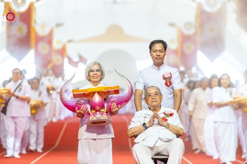 กลุ่มพุทธศาสนิกชนในชุดสีขาวร่วมเดินขบวนในพิธีทอดผ้าป่า โดยมีผู้ถือพานพุ่มผ้าไตรนำหน้า และมีผู้ชายเข็นรถเข็นที่มีชายสูงวัยนั่งพนมมืออยู่ด้านหลัง ภายในสภาธรรมกายสากล
