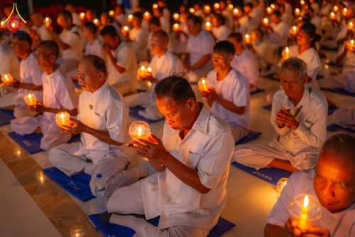 กลุ่มคนสวมชุดขาวนั่งสมาธิถือเทียนในโคมแก้วท่ามกลางแสงสลัว