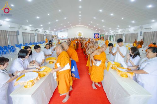 พระภิกษุและผู้เข้าร่วมสัมมนาในชุดขาวกำลังรับเอกสารจากโต๊ะลงทะเบียนภายในห้องประชุมขนาดใหญ่