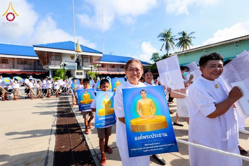 กลุ่มคนในชุดขาวถือป้ายภาพพระประธานสีทองเดินเรียงแถวในพิธีทางศาสนา