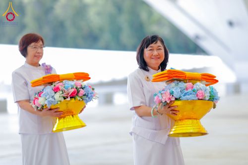 หญิงสองท่านสวมชุดสีขาวถือพานสีทองที่มีดอกไม้และผ้าไตรวางอยู่ด้านบน