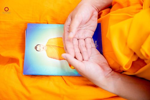 มือของบุคคลในชุดจีวรสีส้มวางอยู่บนหนังสือที่มีรูปภาพพระพุทธรูปบนหน้าปก