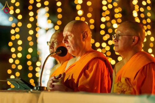 พระภิกษุสงฆ์สามรูปกำลังสนทนาธรรมหน้าไมโครโฟนท่ามกลางแสงไฟประดับในยามค่ำคืน