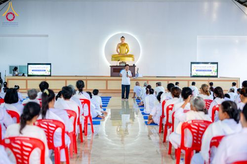 วิทยากรยืนบรรยายธรรมให้ผู้เข้าร่วมโครงการอบรมที่นั่งฟังอยู่ภายในอาคารที่มีพระพุทธรูปเป็นฉากหลัง