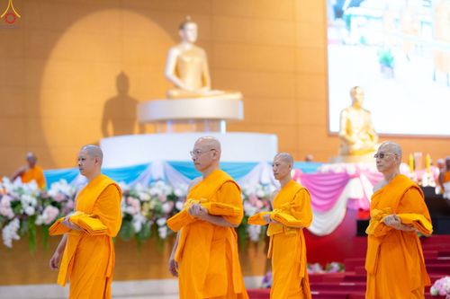 พระภิกษุสงฆ์หลายรูปถือผ้าไตรจีวรเดินในพิธีทางศาสนาภายในอาคารที่มีพระพุทธรูปเป็นฉากหลัง