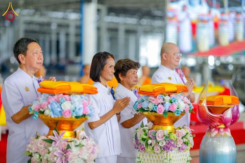 กลุ่มพุทธศาสนิกชนในชุดขาวกำลังยืนพนมมือในพิธีทางศาสนาที่มีพานพุ่มดอกไม้และผ้าไตรวางอยู่ด้านหน้า