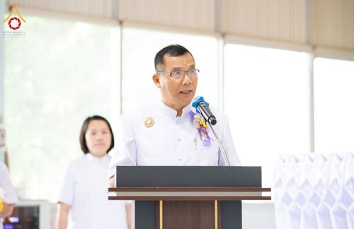 ชายสวมชุดขาวกำลังพูดผ่านไมโครโฟนที่โพเดียมในงานพิธี