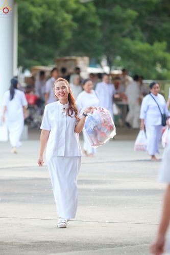 หญิงสาวสวมชุดสีขาวถือถุงของตักบาตรเดินอยู่ในบริเวณวัดที่มีผู้คนสวมชุดขาวเดินอยู่โดยรอบ