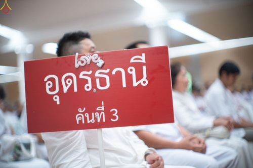 ป้ายสีแดงเขียนว่า อุดรธานี คันที่ 3 ในมือของผู้เข้าร่วมงานที่สวมชุดสีขาว