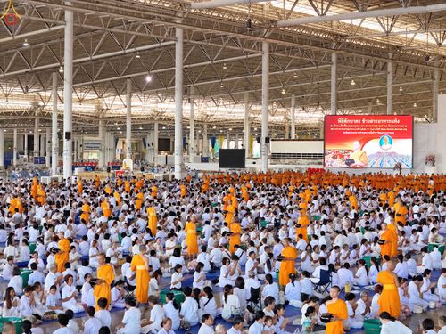 พุทธศาสนิกชนสวมชุดขาวนั่งร่วมพิธีตักบาตรข้าวสารอาหารแห้งภายในอาคารขนาดใหญ่ที่มีพระภิกษุสงฆ์เดินรับบิณฑบาต