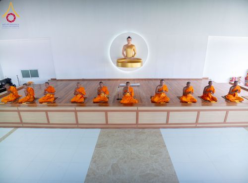พระภิกษุสงฆ์นั่งเรียงแถวบนเวทีไม้เพื่อปฏิบัติธรรมต่อหน้าพระพุทธรูป
