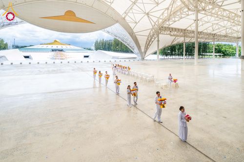 กลุ่มคนชุดขาวถือพานดอกไม้เดินเป็นแถวภายในอาคารสภาธรรมกายสากล