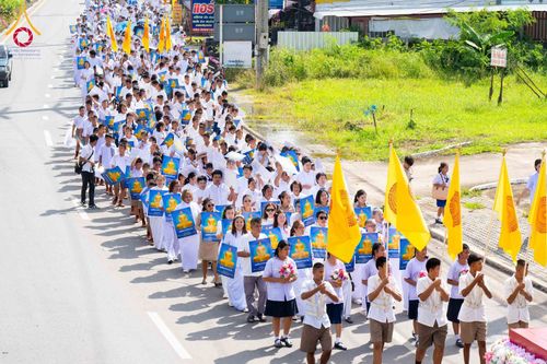 กลุ่มคนสวมชุดสีขาวเดินเรียงแถวบนถนน ถือป้ายภาพพระพุทธรูปและธงสีเหลืองในขบวนพิธีทางศาสนา