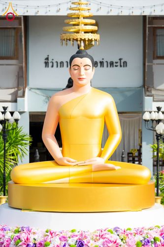 พระพุทธรูปปางสมาธิสีทองประดิษฐานอยู่หน้าอาคาร มีฐานรองรับและประดับด้วยดอกไม้